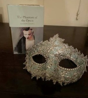 A copy of "The Phantom of the Opera" with a masquerade mask to capture the essence of Phantom.