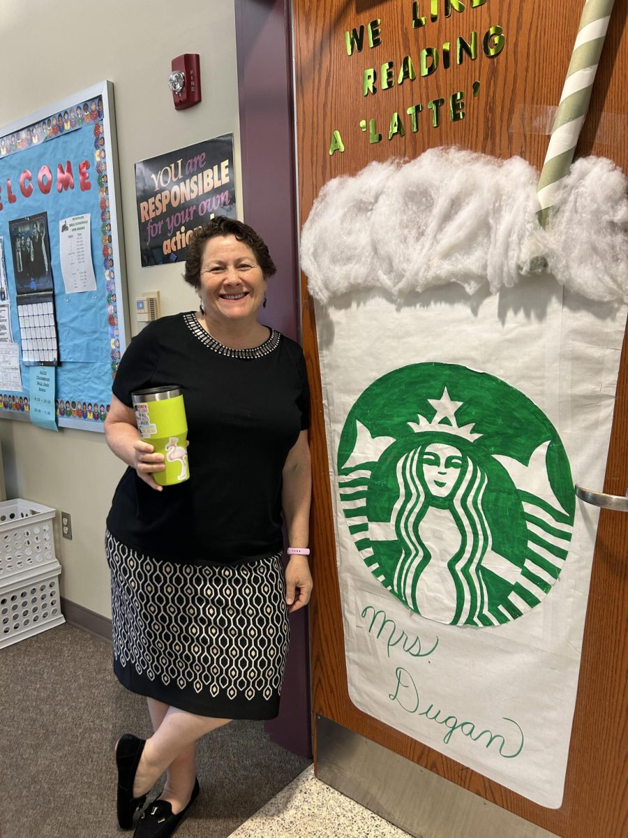 Sixth grade English teacher Michele Dugan posing in front of her coffee themed classroom door with her coffee in hand!
